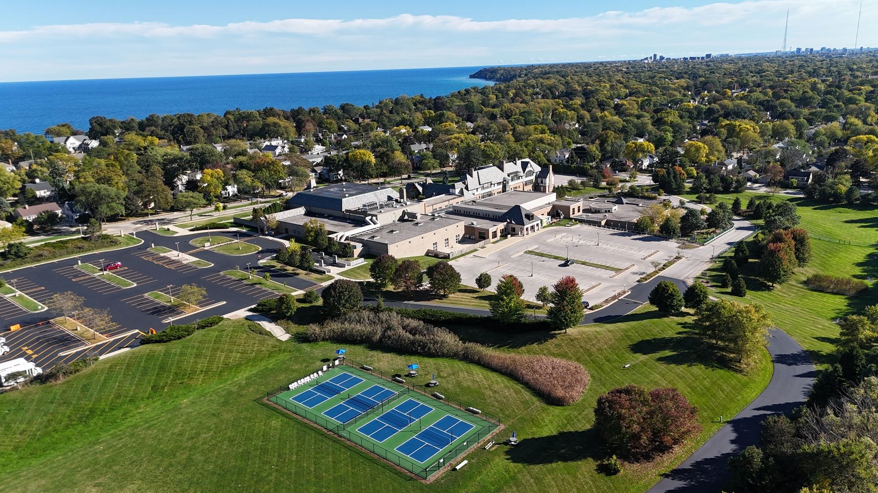 Aerial view of a fenced four-court pickleball facility on a campus lawn with Lake Michigan in the distance