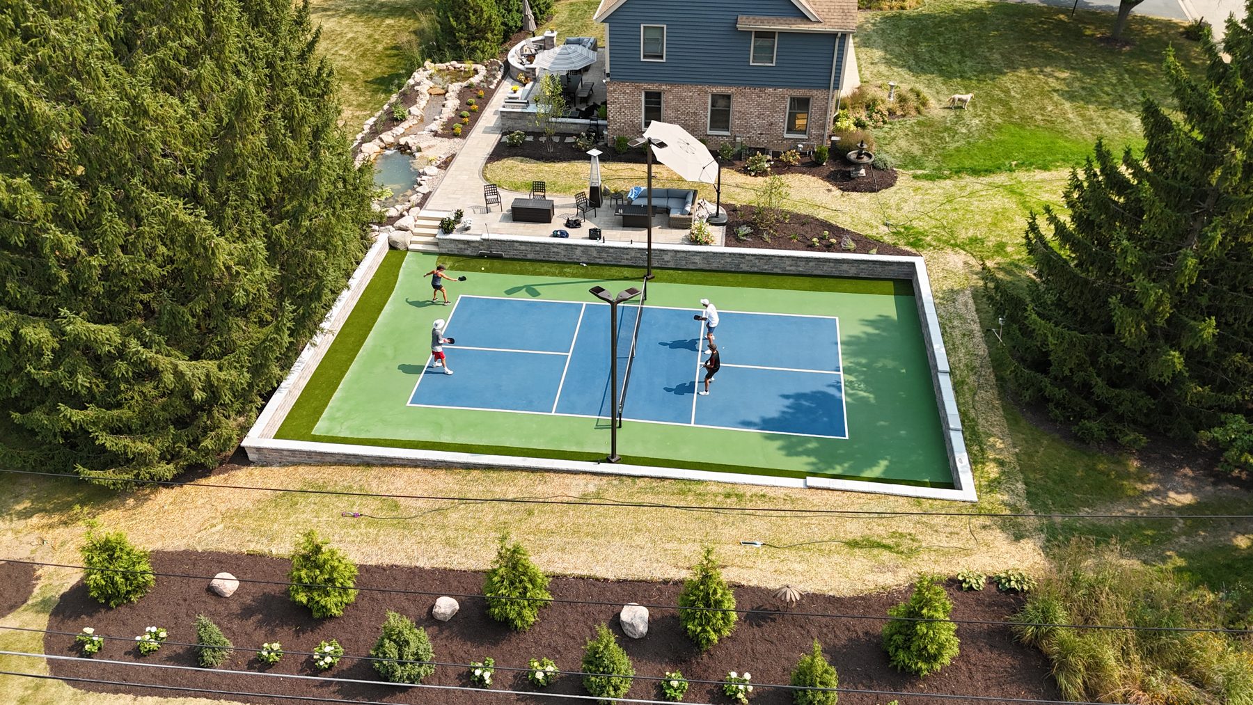 Four players on a blue and green court with a synthetic turf border, stone seat wall, court lights and a patio beside the home